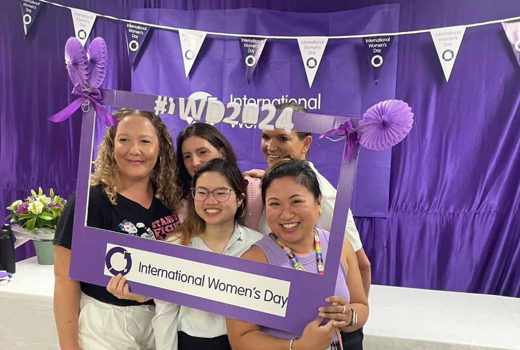 Female Journey Makers (people who work at Transdev) holding up a placard with International Women's Day. They are smiling. In the background, a table with finger food is set up against a backdrop of purple curtains, balloons and International Women's Day branding.
