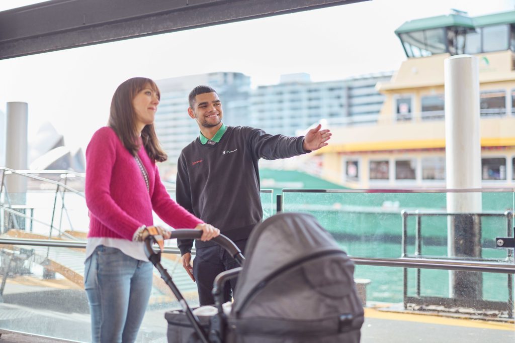 An employee of Transdev Sydney Ferries provides directions to a woman in a pink sweater with a pram. They are at Sydney's Circular Quay Wharf.