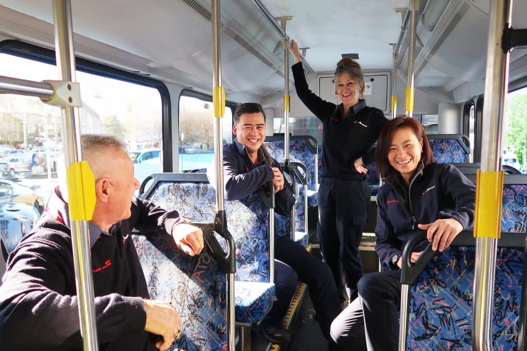 Four bus drivers stand inside a bus and smile at the camera