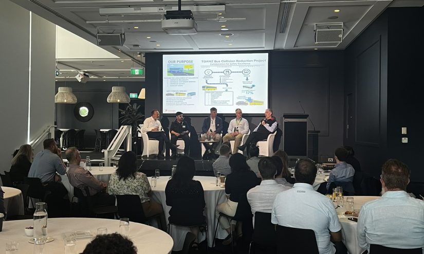 Wide shot of the conference room showing a panel of five speakers on stage discussing safety initiatives while attendees watch from tables in the foreground.